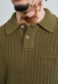 Olive green knitted polo shirt featuring textured open-weave pattern, two brown buttons, and a ribbed collar. Small chest pocket detail.