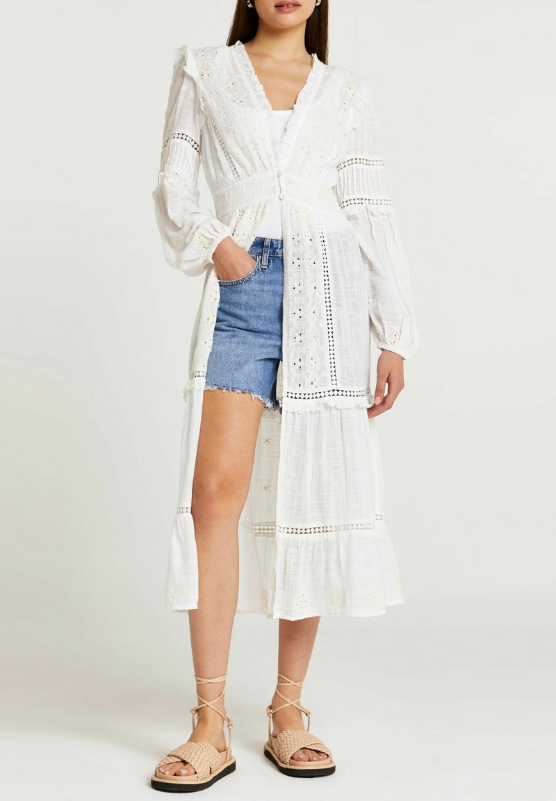 Femme portant un long cardigan en dentelle blanche à œillets sur un top blanc et un short en jean, assorti de sandales beiges tressées avec des liens autour de la cheville.