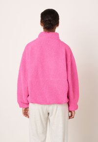 Roza flis pulover z visokim ovratnikom, sproščenega kroja, teksturirane površine in spuščene ramenske linije, v kombinaciji s svetlo sivimi trenirkami.