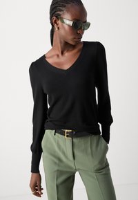 Schwarzer langärmliger V-Ausschnitt-Pullover mit einer weichen Textur, kombiniert mit hellgrünen, maßgeschneiderten Hosen und einem schwarzen Gürtel mit einer goldenen Schnalle.