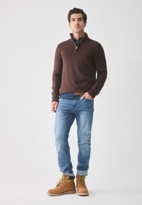 Maglione marrone a maglia con colletto a zip, camicia a quadri blu sotto, jeans azzurri chiari e stivali da lavoro beige con dettagli gialli.