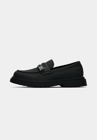 Des mocassins noirs en matériau lisse, avec une sangle logo sur le cou-de-pied, une semelle en caoutchouc épaisse et un design de bout rond.
