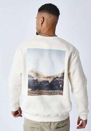 Cream-colored sweatshirt featuring a graphic of rocky mountains and beach on the back. Text reads "PROJECT x PARIS" at the bottom.