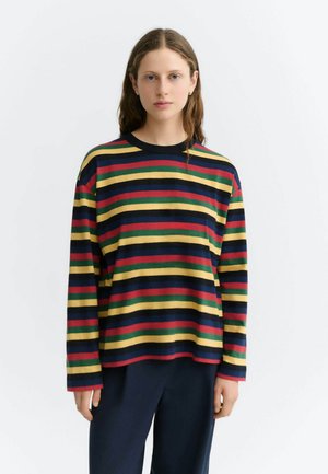 Jeune femme aux longs cheveux portant une chemise à manches longues avec des rayures horizontales rouges, jaunes, vertes et bleu marine et un pantalon foncé.