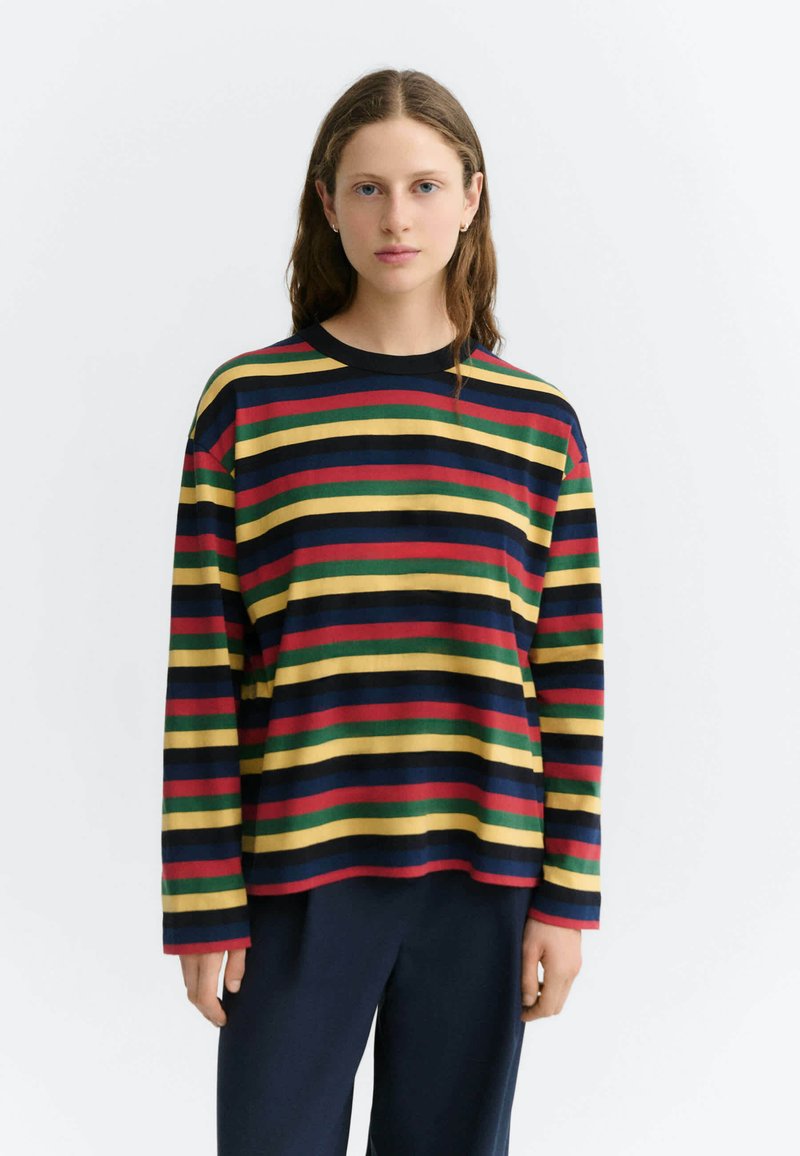 Jeune femme aux longs cheveux portant une chemise à manches longues avec des rayures horizontales rouges, jaunes, vertes et bleu marine et un pantalon foncé.
