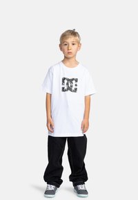 Camiseta blanca de manga corta con un logo gráfico negro, combinada con pantalones negros holgados y zapatos grises con cordones blancos y suelas turquesas.