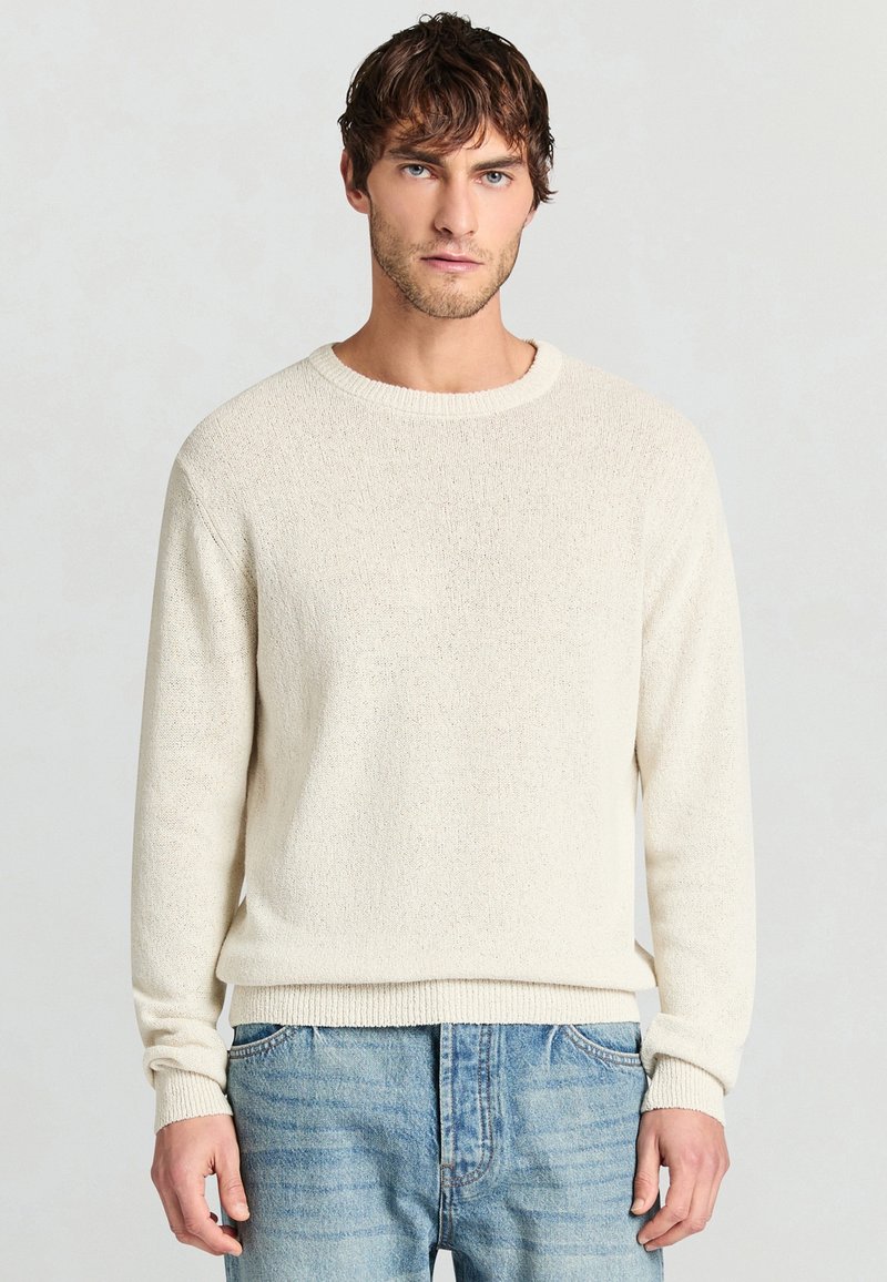 Mann mit kurzen braunen Haaren, der einen strukturierten, cremeweißen Pullover und hellblaue Jeans trägt, vor einem schlichten Hintergrund stehend.