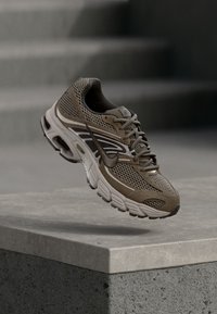 Καφέ αθλητικό παπούτσι με επάνω μέρος από δίχτυ, ανοιχτό μπεζ λεπτομέρειες, στρογγυλεμένη μύτη και μαλακή σόλα, εκτεθειμένο σε τσιμεντένιο βάθρο.