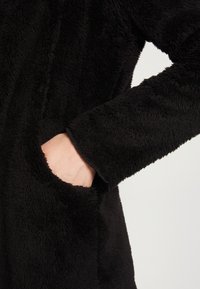 Zwarte fleecejas met een zachte, pluche textuur. Voorzien van een zijzak en een brede mouwopening. Gladde stiksels langs de randen.