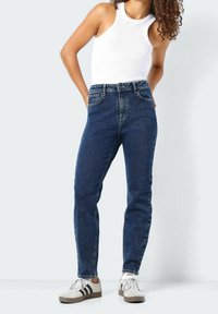 Džínové kalhoty s vysokým pasem v modré barvě s klasickým střihem, pěti kapsami a minimálním opotřebením, v kombinaci s bílým tílkem a teniskami.