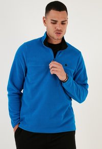 Pullover in pile blu con colletto a mezza cerniera, dettagli neri sul collo e un piccolo logo sul lato sinistro del petto. Tessuto morbido e maniche lunghe.