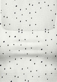 Tissu blanc avec de petites taches noires irrégulières dispersées et une texture côtelée subtile, montré en gros plan.