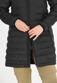 Sort pufferjakke med lodret kviltet design, lynlås-lukning og sidelommer. Modellen justerer lynlåsen, mens han har khaki bukser på.