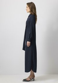 Rochie bleu marin cu dungi, guler, mâneci lungi și slituri laterale. Materialul are o textură netedă; asortată cu pantofi negri cu vârf ascuțit.