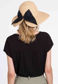 Breitkrempiger Strohhut mit einer schwarzen Stoffschleife hinten. Natürliche Farbe mit strukturiertem Finish. Getragen mit einem schwarzen Hemd und beigen Hosen.