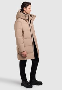 Hombre de pie de lado, vistiendo un abrigo de invierno acolchado con capucha beige, pantalones negros y botas gruesas negras, contra un fondo liso.