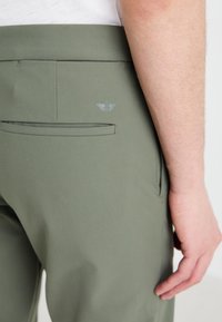 Olive green pants made from smooth fabric, featuring a back pocket and a subtle logo above it. Simple, tailored design with clean lines.