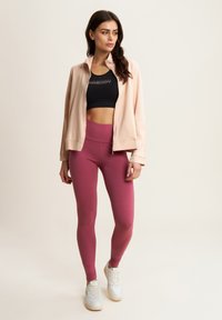 Giacca zip con cappuccio rosa chiaro sopra un crop top nero e leggings bordeaux a vita alta, abbinata a sneakers bianche. Texture lisce dappertutto.