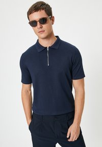 Mörkblå kortärmad polo med dragkedja i kragen, ribbad struktur och modern passform, matchad med mörkblå byxor. Tillbehör: solglasögon.