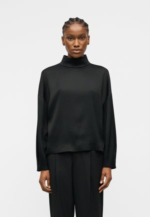 Femme aux cheveux tressés portant un pull à col roulé noir ample et un pantalon noir, debout devant un fond blanc uni.