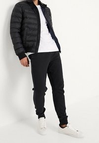 Doudoune noire avec fermeture éclair à l'avant et poignets côtelés, associée à un pantalon de survêtement noir à coupe ajustée et des sneakers blanches.