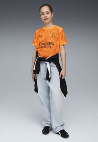 Maglia arancione brillante a maniche corte con motivi astratti, logo nero e testo. Abbinata a jeans ampi blu chiaro e scarpe nere.