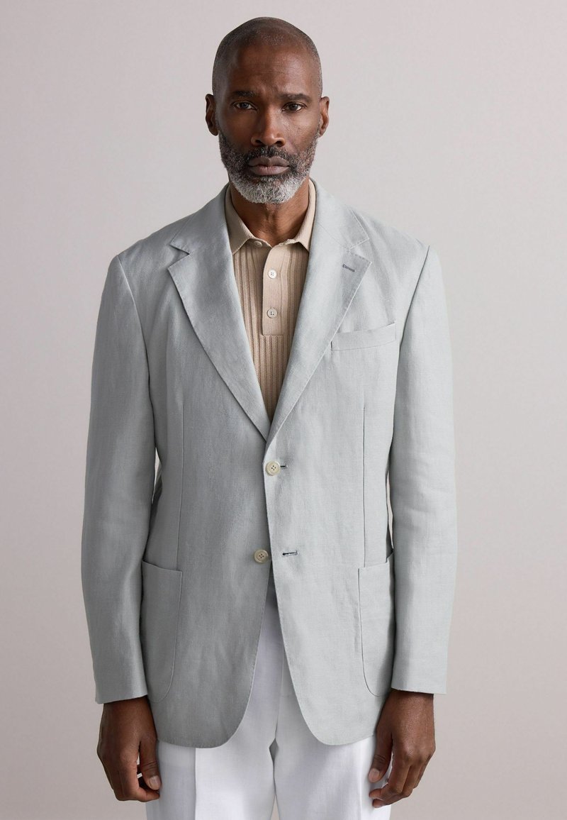 Blazer en lin gris clair avec revers cranté, deux poches avant, et chemise beige à boutons en dessous. Silhouette simple et ajustée.