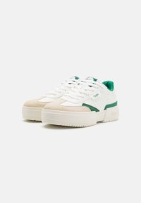 Baskets blanches et beiges avec des accents verts. Caractérisées par des matériaux synthétiques lisses et en suède, un design à lacets et une semelle épaisse en caoutchouc.