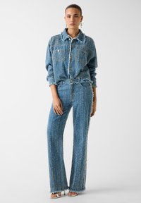 Completo giacca e pantaloni in denim blu, caratterizzato da una finitura strutturata con orli sfrangiati, grandi tasche e bottoni dorati. Vestibilità su misura.