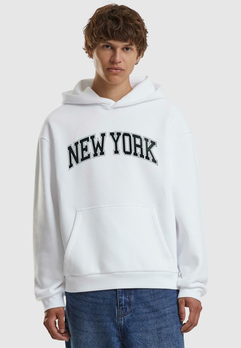 Biela mikina s kapucňou s nápisom "NEW YORK" v čiernom písme, predné klokanie vrecko a rebrované manžety a lem. Nosí sa s modrými džínsami.