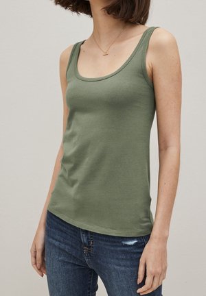 Femme portant un débardeur ajusté vert olive et un jean bleu, avec un collier en or délicat, se tenant devant un fond clair.