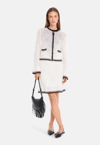 Weißer gestrickter Blazer mit schwarzen Akzenten, kombiniert mit einem passenden Kleid und einer schwarzen Fransen-Handtasche. Schwarze spitze Ballerinas runden den Look ab.