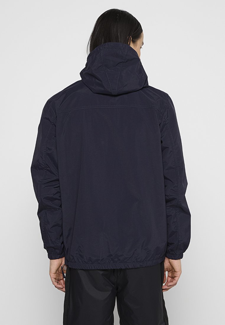 Chaqueta con capucha de color navy con puños y dobladillo elásticos. Material ligero, con una textura suave y un detalle de costura mínima en la parte posterior.