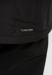 Svart climastorm-jacka med texturerad yta och kontrasterande vitt logotyp på nederkanten. Har en slank och funktionell design.