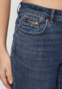 Mørkeblå denim jeans med et nærbillede af en sidelomme, der har en metallisk lynlås og kontrasterende syninger, der fremhæver en glat tekstur.