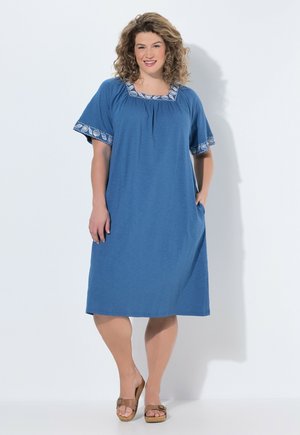 Kobieta o kręconych włosach w niebieskiej sukience do kolan z ozdobnym wzorem na rękawach i dekolcie, stojąca z jedną ręką w kieszeni, uśmiechająca się.