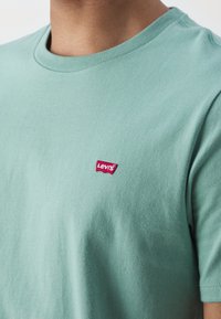 Gros plan d'une personne portant un t-shirt vert menthe Levi's avec un petit patch rouge du logo Levi's sur la poitrine gauche.