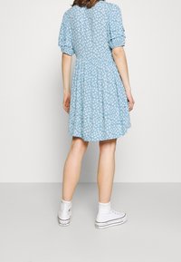 Abito azzurro chiaro con motivi floreali bianchi, maniche a sbuffo corte e gonna svasata. Indossato con sneakers bianche alte.