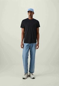 Man som bär en svart t-shirt, ljusblå jeans, grå sneakers och en ljusblå basebollkeps, stående mot en enfärgad ljus bakgrund.