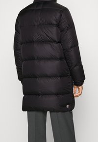 Puffjacka i svart, med quiltade sektioner, hög krage och en logotypplatta nära fållen. Tillverkad i glänsande, slätt material.