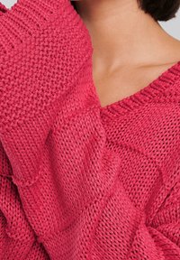 Strickpullover in lebhaftem Pink. Mit V-Ausschnitt, strukturiertem Muster und lockeren Ärmeln. Das Material scheint weich und atmungsaktiv zu sein.