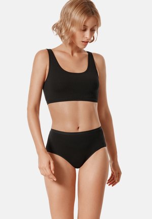 Woman with short blonde hair wearing a black sports bra and high-waisted underwear standing against a plain light background.
