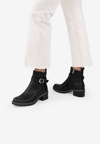 Stivali alla caviglia in pelle nera con punta arrotondata, cerniera laterale e dettaglio con fibbia; tacco basso e texture liscia. Gambe dei pantaloni di colore chiaro visibili.