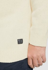 Beige gebreide trui met een geribbelde manchette, voorzien van een klein zwart label met de tekst "s.O" nabij de zoom. De textuur lijkt zacht en casual.