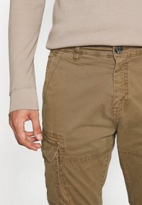 Khaki spodnie cargo o gładkiej fakturze, z zapięciem na guzik z przodu, bocznymi kieszeniami i subtelnymi detalami w szyciu. W połączeniu z jasnobeżową koszulą z długim rękawem.