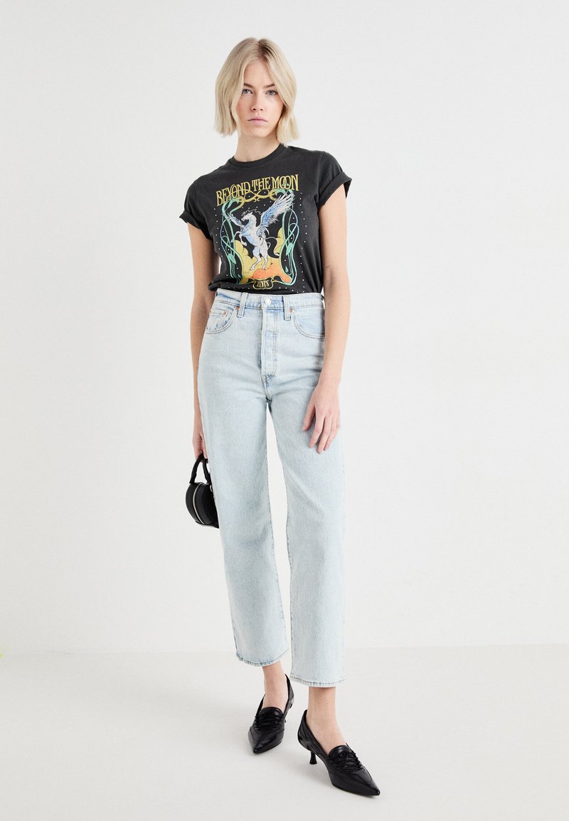 Camiseta negra gráfica con un diseño mitológico, jeans rectos de lavado claro, zapatos de tacón negros y un pequeño bolso negro.