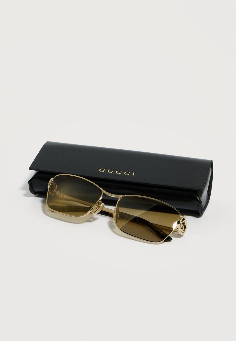 Lunettes de soleil rectangulaires à monture dorée avec lentilles marron reposant devant un étui Gucci noir sur une surface blanche.