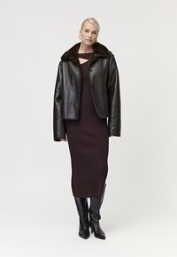 Femme portant une veste en cuir noire avec col en fourrure, une robe midi côtelée foncée, des bottines noires à talons et des boucles d'oreilles créoles dorées, debout sur un fond blanc.