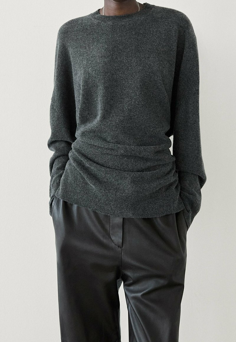 Pull gris foncé en maille avec un col rond et des détails froncés à la taille, associé à un pantalon en cuir noir. Texture douce et coupe ample.