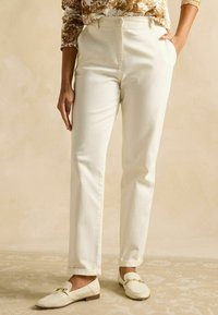 Pantaloni croiți, de culoare crem, cu croială dreaptă, cu buzunare laterale și închidere cu nasture, asortați cu pantofi loafer de aceeași culoare.